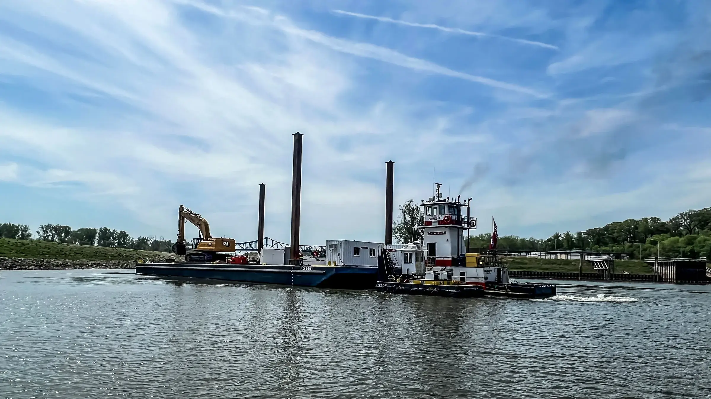 A barge operates on a river with an excavator aboard