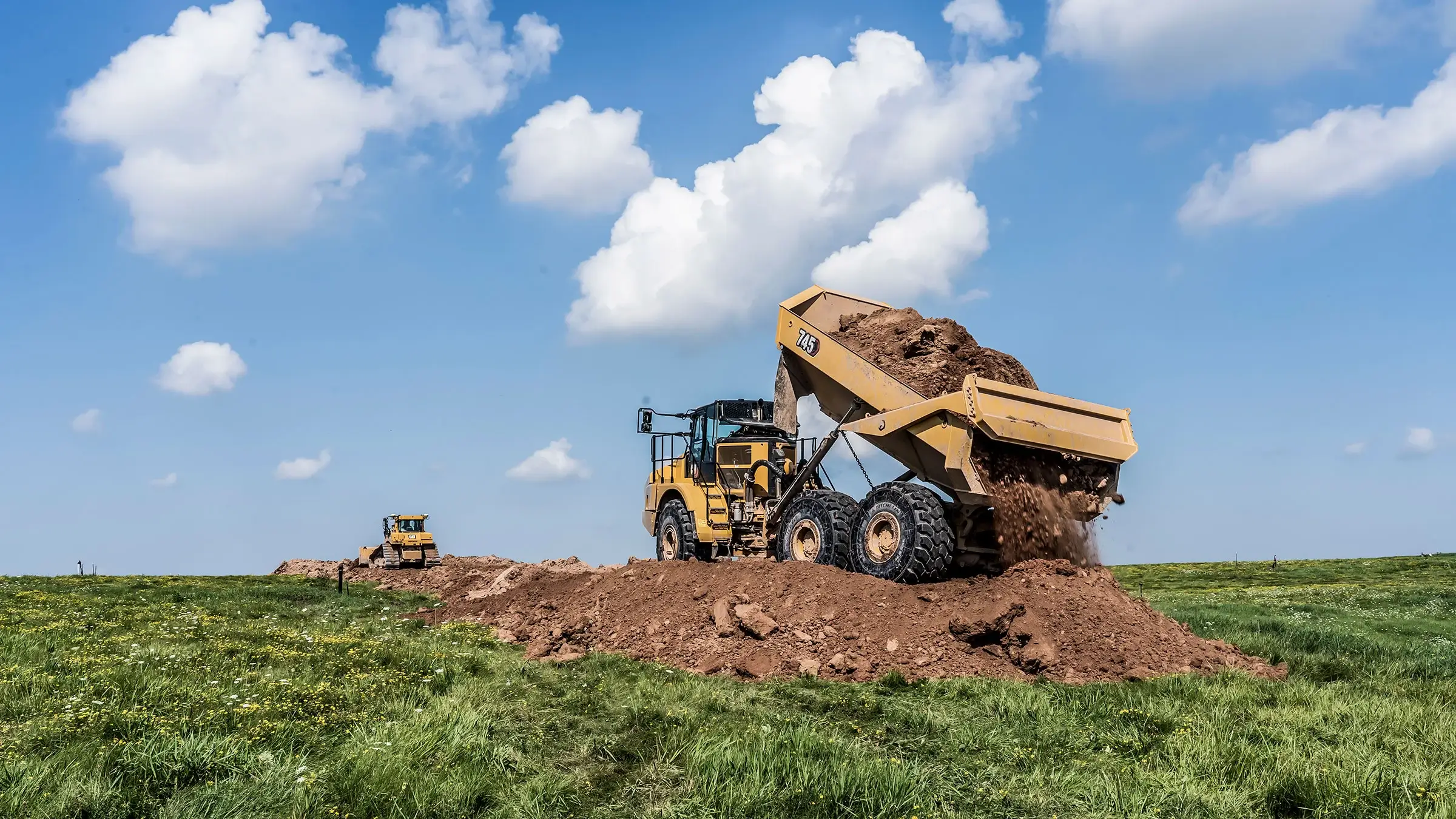 A large dump truck dumps dirt in a pile in a grassy field