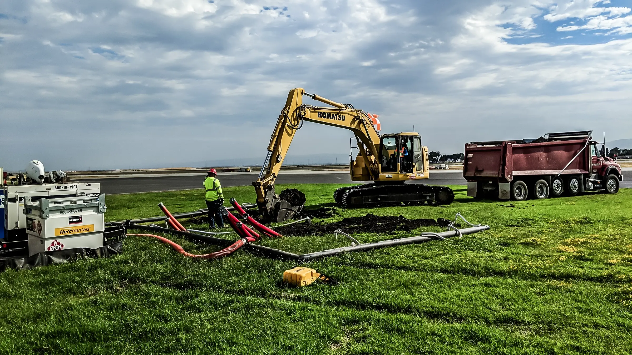 An excavator and crew member work to insert cable into ground near airfield