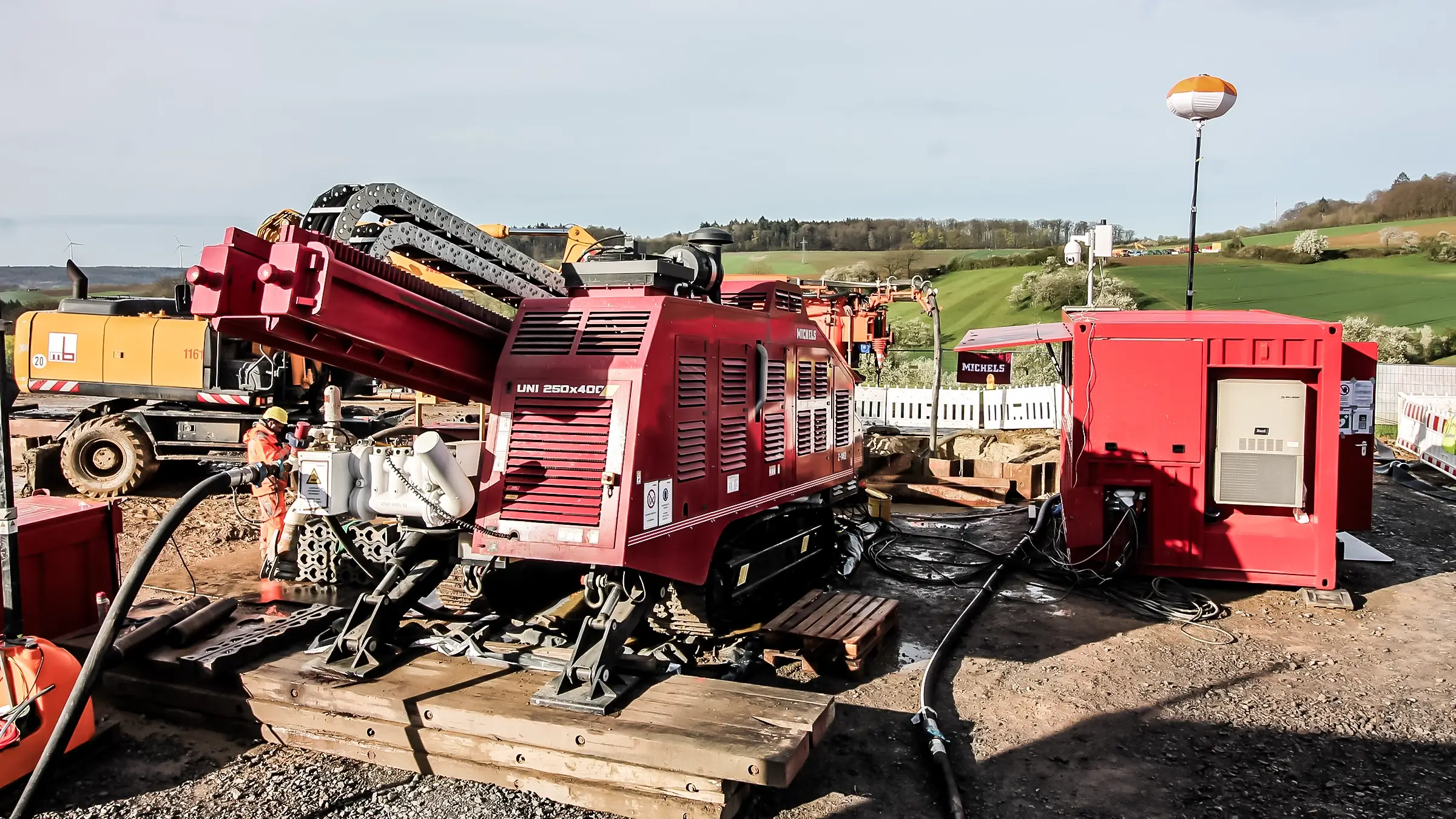 An HDD rig operating with an excavator in a rural hilly area.
