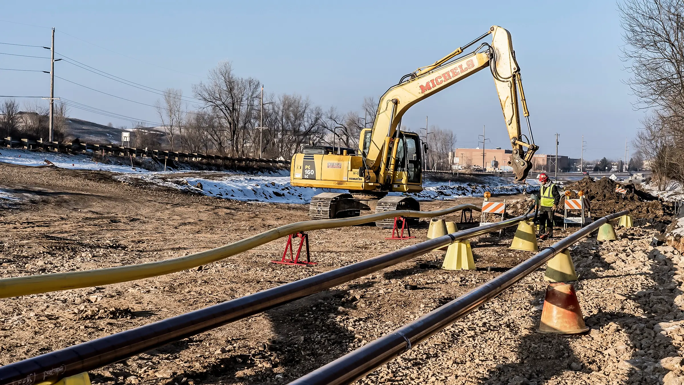 An excavator operates near two pipelines in a rural snowy environment.