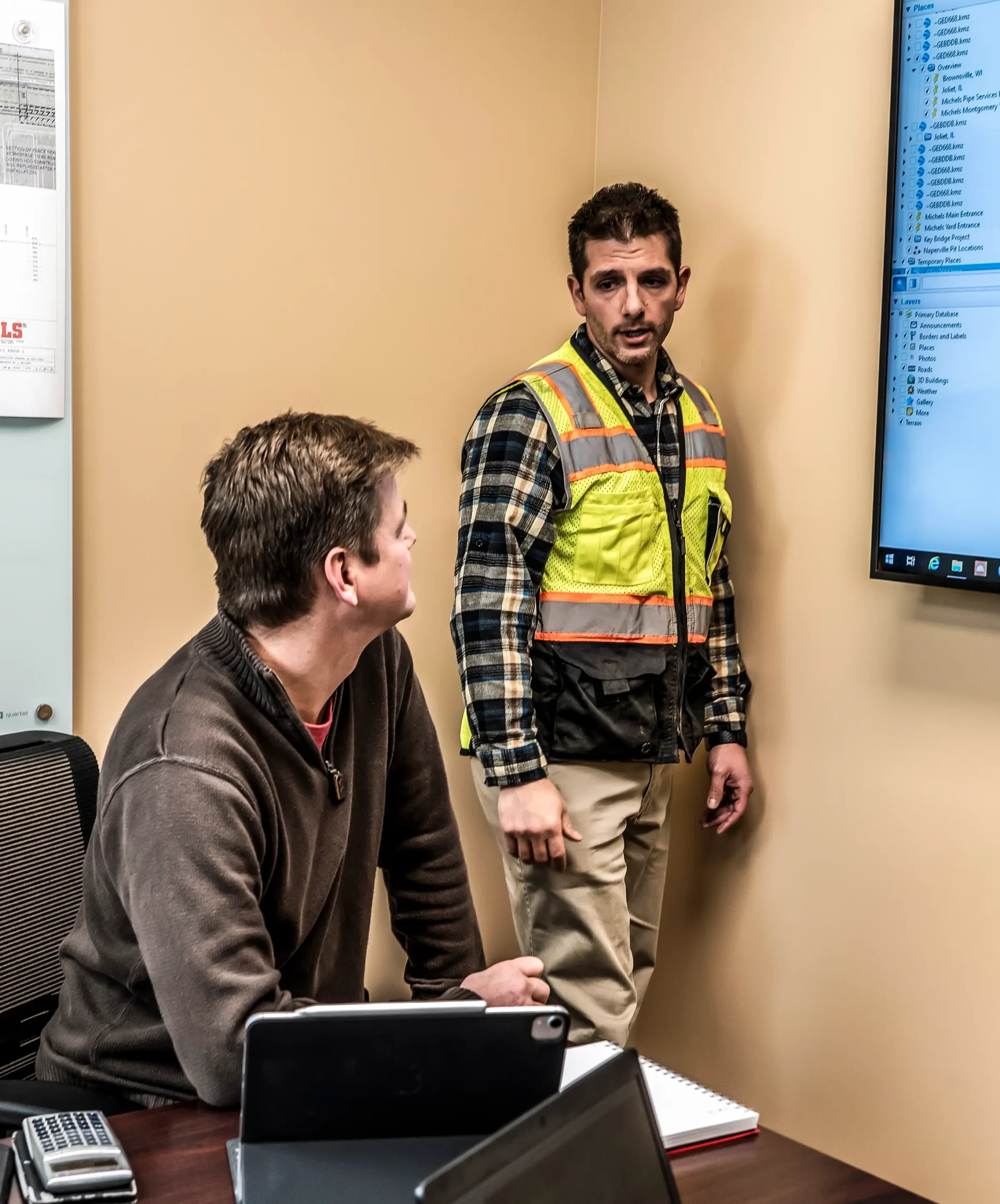 One man wearing a reflective vest lectures another in a conference room.